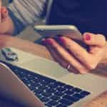 A person sitting at a desk in front of a laptop while holding a smartphone in their left hand.