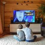 Two children sitting side by side while watching a cartoon on the television in a wooden living room.