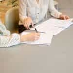 Two women sitting at a table with documents with one of them signing on them.