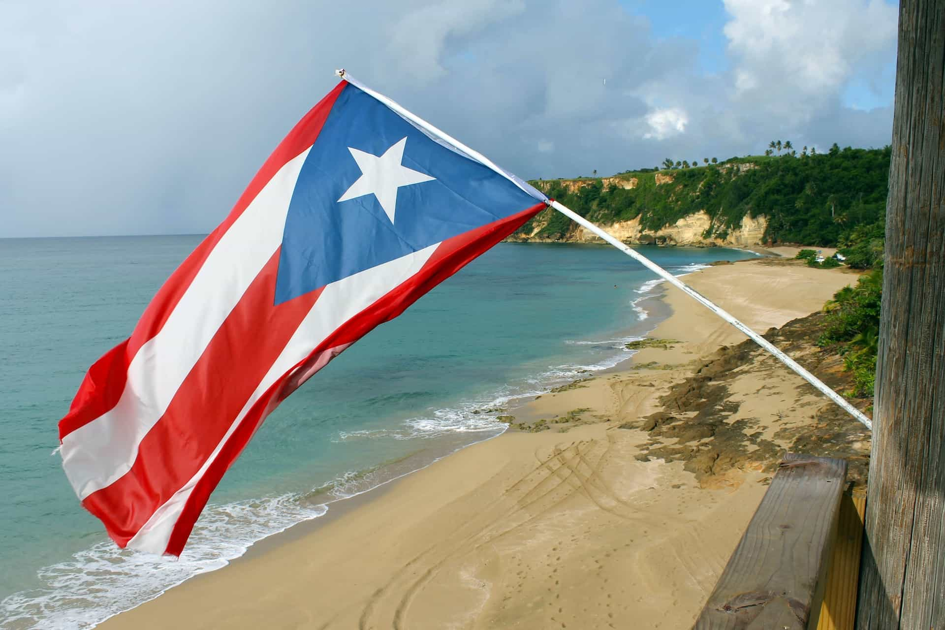 The flag of Puerto Rico flying above a sandy beach, with tree-covered cliffs and the blue ocean extending out behind it.