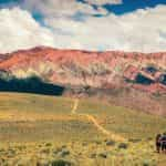 A couple walks up a hill covered in green and yellow shrubs at the base of red mountains in Jujuy, Argentina.