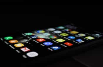 A smartphone lying flat on its back in darkness while displaying its home screen with many different mobile app icons on it.